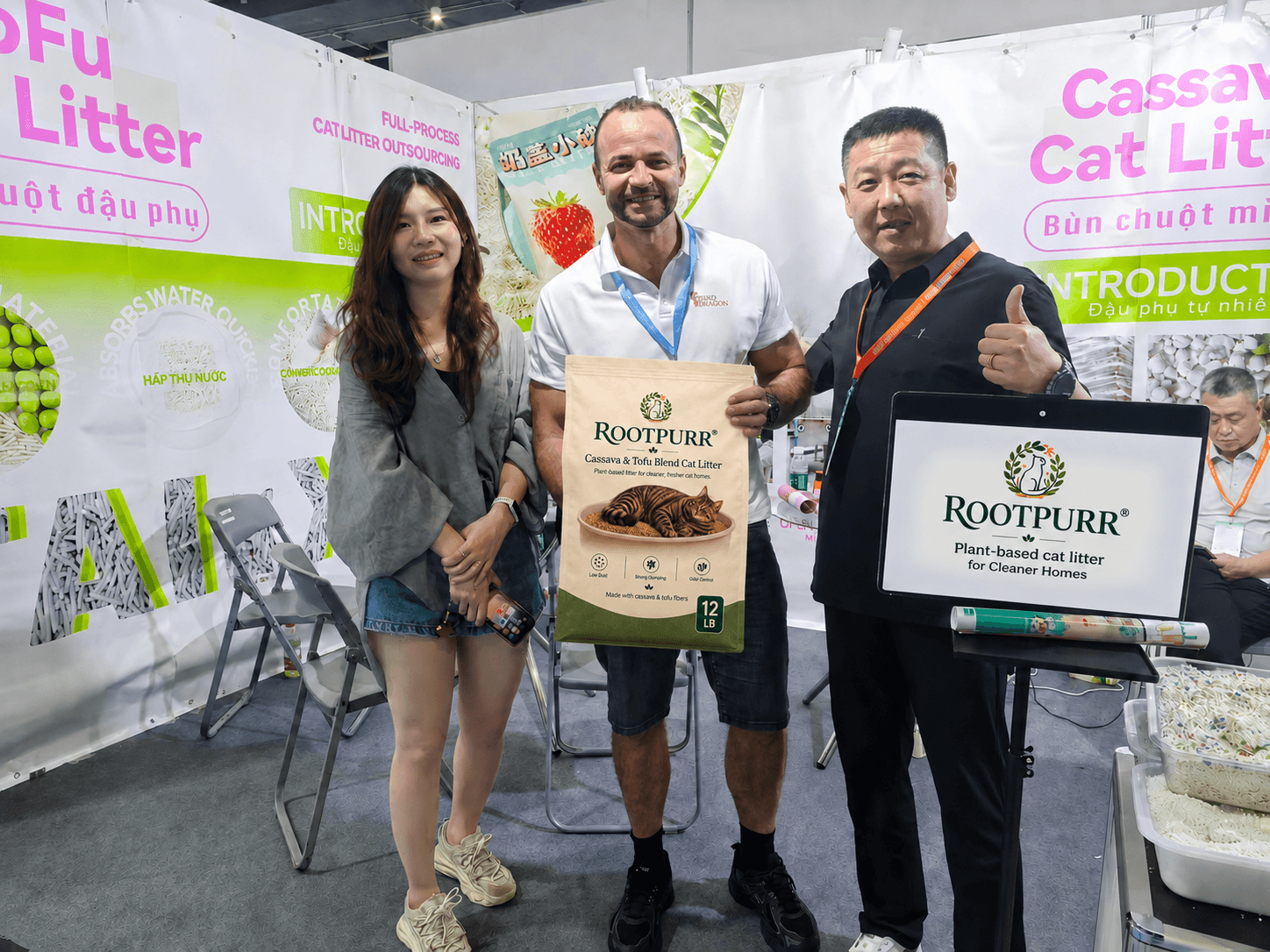 Three people at the RootPurr trade-show booth holding a RootPurr Cassava & Tofu Blend 12 lb cat litter bag in front of the Cassava Cat Litter backdrop.