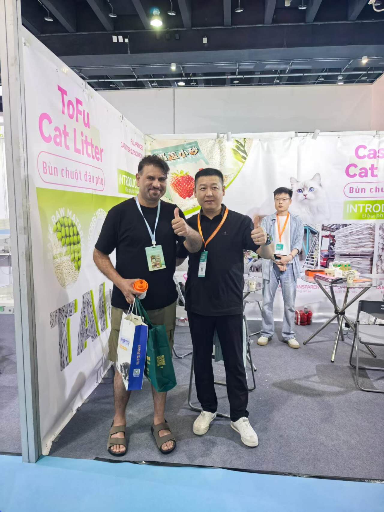 Two buyers giving a thumbs-up at the RootPurr ToFu Cat Litter trade-show booth, holding a RootPurr gift bag.
